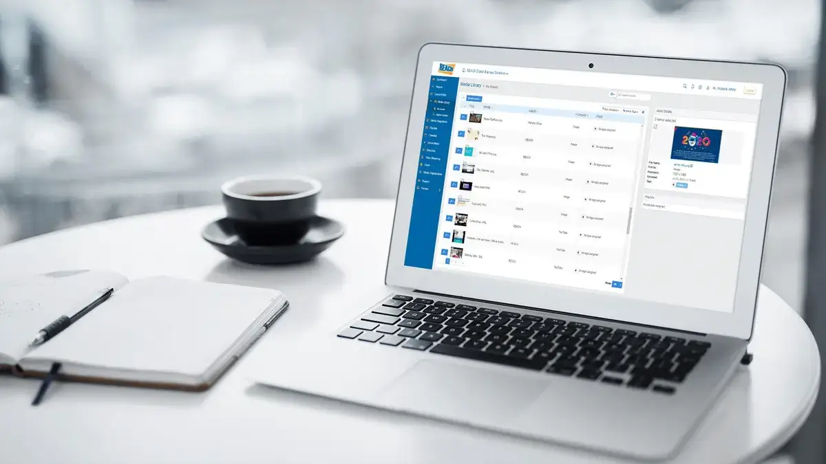 REACH Media Network digital CMS software featured on work laptop, on table with cup of coffee and notebook.